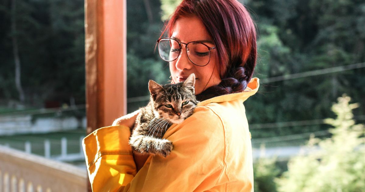 woman with glasses and in a yellow jacket stands holding a tabby cat in her arms, and smiling down at it